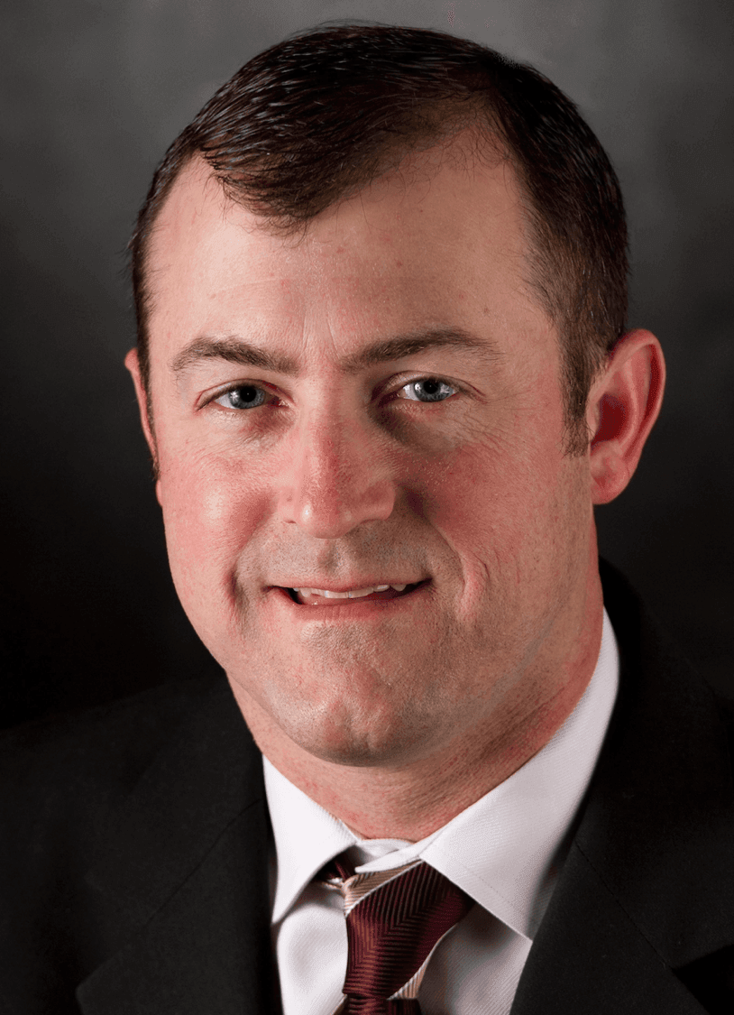 Close-up portrait of a man in a suit and tie, smiling against a dark background.