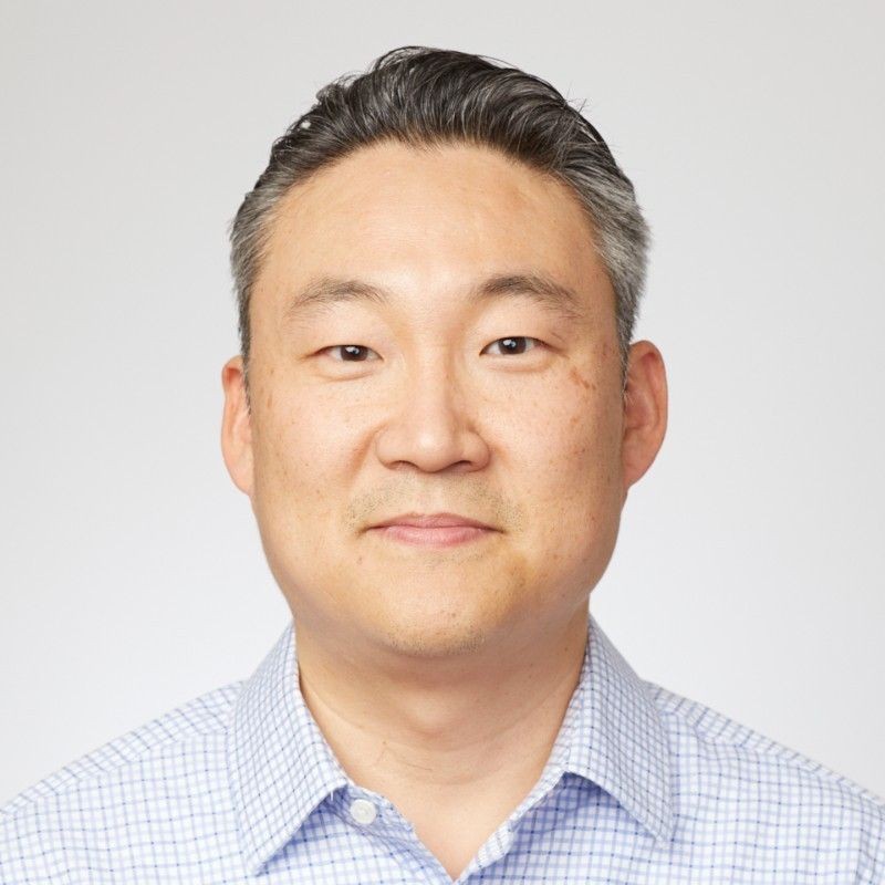 Portrait of a man with short hair wearing a checked shirt, smiling slightly against a plain background.